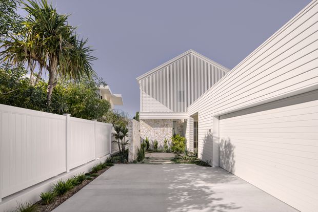 A bright residential driveway and entry approach, framed by a white horizontal-clad garage and a taller gabled volume with vertical white cladding. A large white garage door occupies the right side, while a white privacy fence, paved driveway, landscaped planting, and a stone-clad recessed entry create a clean contemporary coastal composition.