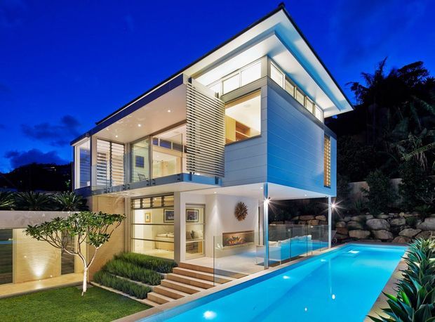 Twilight exterior view of a contemporary multi-level house with crisp white and light-grey cladding, extensive floor-to-ceiling glazing, projecting balconies, horizontal privacy screens, and a flat or low-pitched roof. A narrow illuminated swimming pool runs alongside the building, with landscaped planting, stonework, and exterior stairs visible.