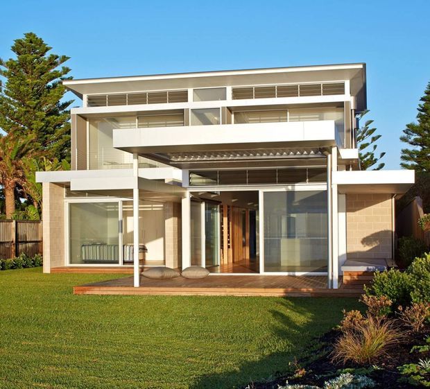 Exterior view of a contemporary multi-storey residence with strong horizontal lines, extensive glazing, white rendered projecting balconies, a deep covered terrace, and warm timber decking. The house is set within a landscaped suburban garden with lawn and mature trees.