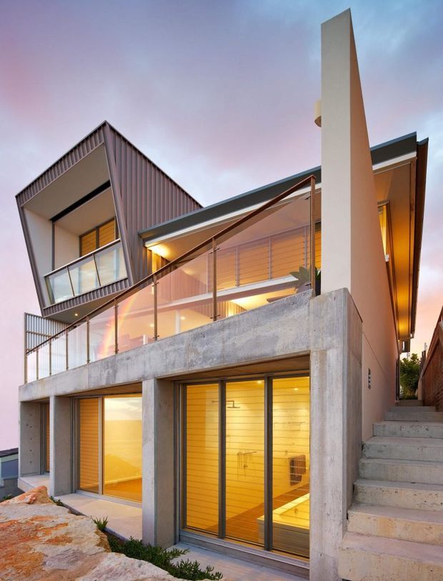 Exterior view of a contemporary multi-level residential building on a sloped site. The house features exposed concrete walls and terraces, dark metal cladding and roof elements, extensive glazing with glass balustrades, warm timber-lined interiors visible through the windows, and a prominent vertical rendered wall beside an external stairway. The image is taken at dusk with warm interior lighting and a pink-blue sky.