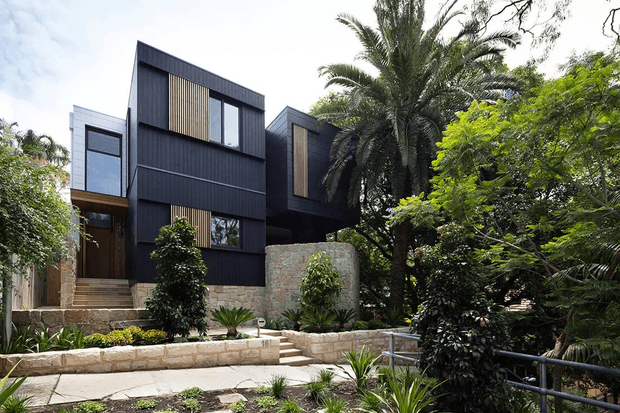 Exterior view of a contemporary multi-level residence set within dense tropical landscaping. The house features dark vertical cladding, projecting box-like volumes, large glazed openings, warm timber screening, and a pale stone retaining wall. A stepped stone-paved path and landscaped front garden lead toward the entrance.