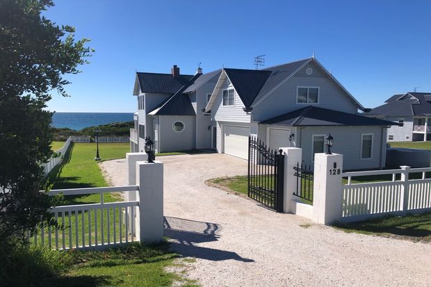 Exterior view of a large coastal-style residential house with multiple steep gables, grey weatherboard cladding, dark roofing, white trim, and a gated gravel driveway. The property overlooks the ocean, with formal fencing, lawn, and an attached garage visible.