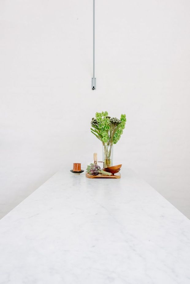 Minimalist interior vignette featuring a long white marble or stone island/tabletop extending toward the camera, against a plain white wall. A small arrangement of greenery in a clear vase, wooden serving pieces, and a small cup sit at the far end. A simple exposed hanging cord is visible above.