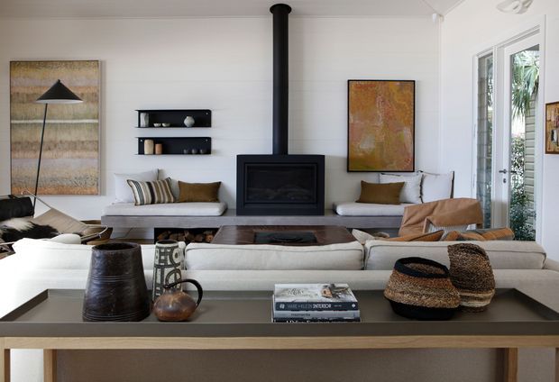 A bright, refined living room with white paneled walls, a central black fireplace and flue, built-in open shelving, pale upholstered sofas, timber furniture, woven accessories, and large framed artwork. Full-height glazed doors open toward greenery, bringing natural light into the room.
