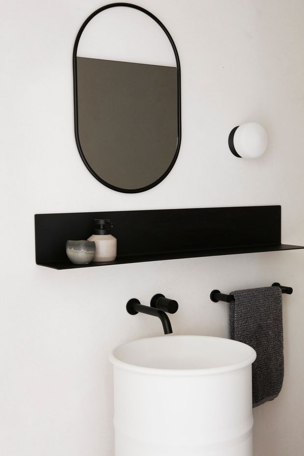 A minimalist bathroom vignette featuring a white cylindrical pedestal basin, matte black wall-mounted tapware, a black shelf with toiletries, a rounded black-framed mirror, a spherical wall light, and a dark grey hand towel. The space has a clean, monochromatic contemporary aesthetic with strong black-and-white contrast.