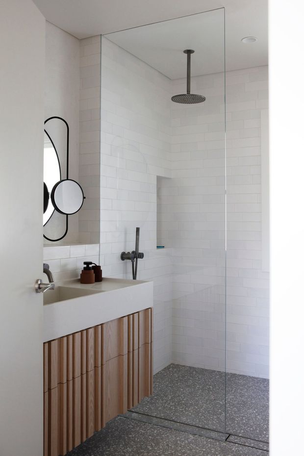 Bright contemporary bathroom featuring a walk-in shower enclosed by a frameless glass screen, white subway-tile walls, a grey terrazzo-style floor, and a floating vanity with a white basin and vertically fluted timber cabinetry. A round black-framed mirror and dark shower fittings provide contrast.