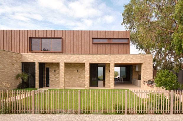 Front exterior view of a contemporary two-storey residence with a warm pale brick lower level and vertically ribbed terracotta-toned cladding to the upper storey. Dark-framed windows and doors open onto a small paved terrace and lawn, enclosed by a vertical timber picket fence. A covered outdoor area and barbecue are visible at the right, with mature planting around the property.