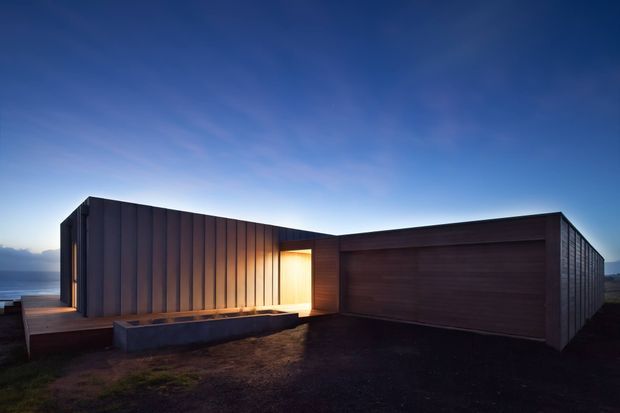A low, angular modern home photographed at dusk, with dark vertical metal cladding, a warm illuminated recessed entry, and a broad dark timber-clad garage or wall volume. The building sits on a coastal or elevated site, with ocean visible to the left and a dramatic blue evening sky.