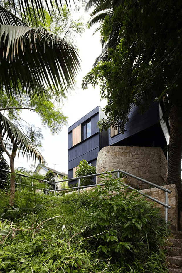 Exterior view of a contemporary multi-level house set within dense tropical landscaping. The dark vertically clad upper structure includes large windows, while a rounded textured natural-stone wall forms the lower level. A planted sloping garden with metal handrails leads up toward the building.