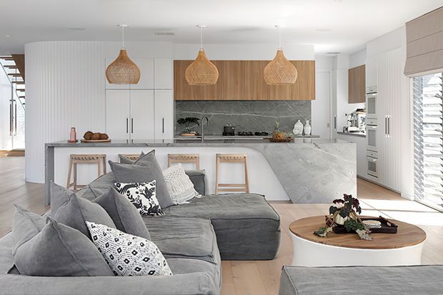 Bright open-plan contemporary living and kitchen area with a large grey sectional sofa in the foreground, a white island with timber stools, integrated white cabinetry, warm timber overhead cabinets, marble-look surfaces, and three woven pendant lights. The space includes a dining or living zone extending toward the kitchen, with abundant natural light and louvred glazing at right.