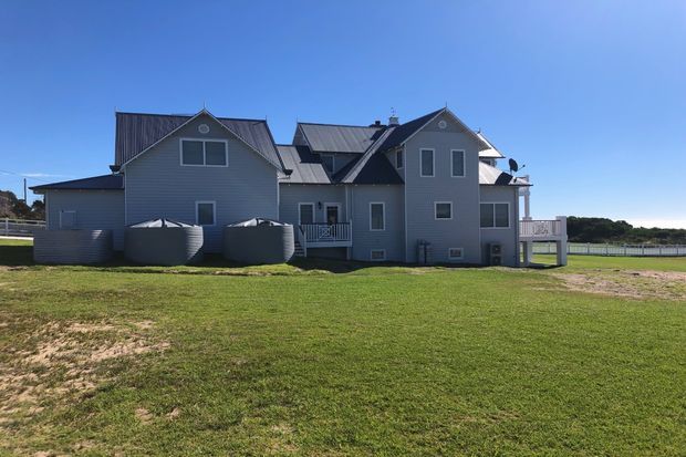 Exterior rear or side view of a large multi-level residential house with grey weatherboard cladding, multiple pitched gables, dark metal roofing, white-trimmed windows, balconies, and large cylindrical water tanks. The home sits on a broad grassy site beside a visible body of water under a clear blue sky.
