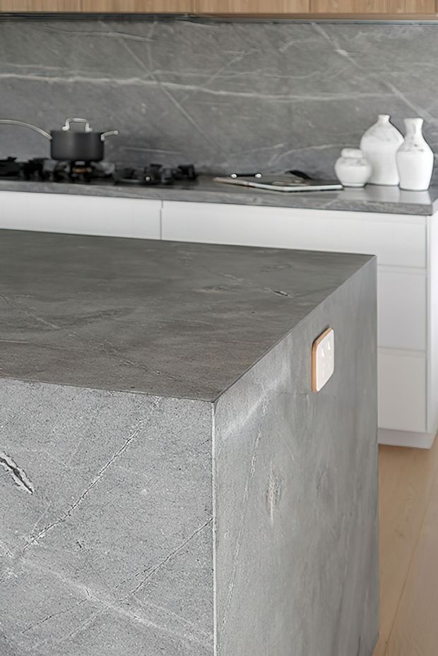 Close-up view of a contemporary kitchen dominated by a large grey stone-finished island with a thick waterfall edge and integrated power outlet. Matching grey stone surfaces form the benchtop and splashback behind a gas cooktop, with white lower cabinetry, timber upper cabinetry, and pale timber flooring. Minimal white ceramic vessels provide restrained styling.