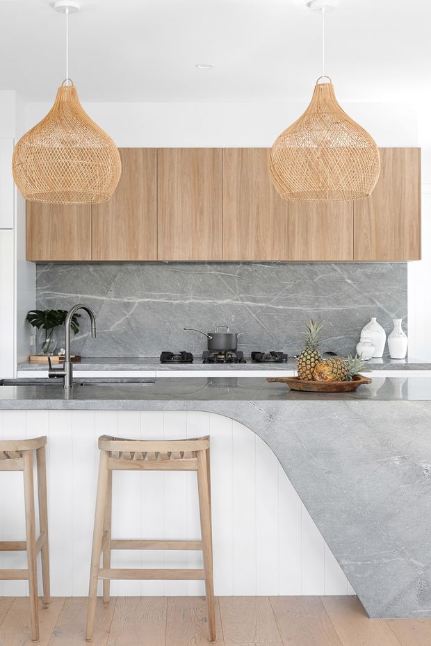 Bright contemporary kitchen featuring full-height light timber cabinetry, a grey veined stone splashback and waterfall island benchtop, white vertical-panel island detailing, timber bar stools, and two oversized woven pendant lights.