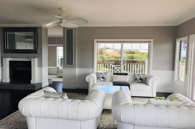 Bright, spacious living room with white tufted sofas and armchairs arranged around a light coffee table, a dark fireplace with ornate mirror, grey walls with white wainscoting and trim, and large windows overlooking a balcony and greenery. A ceiling fan and patterned rug complete the room.