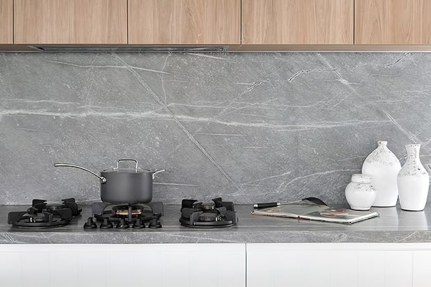 Close-up view of a contemporary kitchen cooking zone with a dark grey veined stone backsplash and matching benchtop, four-burner gas cooktop, dark saucepan, light timber overhead cabinetry, white lower cabinetry, and decorative white ceramic vessels.