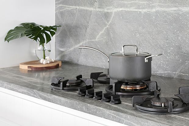 Close-up of a contemporary kitchen cooktop with a grey stone benchtop and matching full-height splashback. A dark grey lidded saucepan sits on one of several black gas burners, with a small potted tropical plant on a wooden tray at the rear.