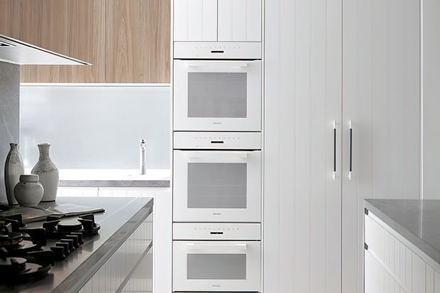 Bright contemporary white kitchen with a bank of integrated stainless-steel ovens, tall white cabinetry, a gas cooktop on a grey island, and light timber upper cabinetry. The space has clean vertical panel detailing and a minimal, refined appearance.