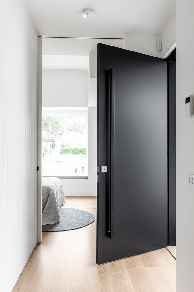 A bright minimalist interior view from a hallway through an open matte-black door into a bedroom. The space features pale timber flooring, white walls and ceiling, a large window, and glimpses of a neatly made bed with a round grey rug. The black door and hardware provide a strong graphic contrast against the light finishes.