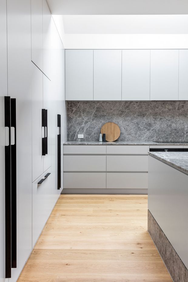 A refined minimalist kitchen with full-height pale cabinetry, integrated black vertical handles, a grey stone benchtop and matching stone splashback, and light timber flooring. A central island extends into the foreground, while a skylight or recessed ceiling opening brings soft natural light into the space.