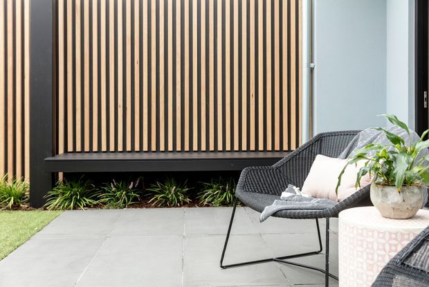 A contemporary outdoor courtyard or patio featuring large grey paving slabs, a black metal-framed timber vertical-slat privacy screen, low ornamental planting, and woven outdoor lounge chairs with a pale patterned side table and potted greenery. The space has a clean, relaxed, modern character.
