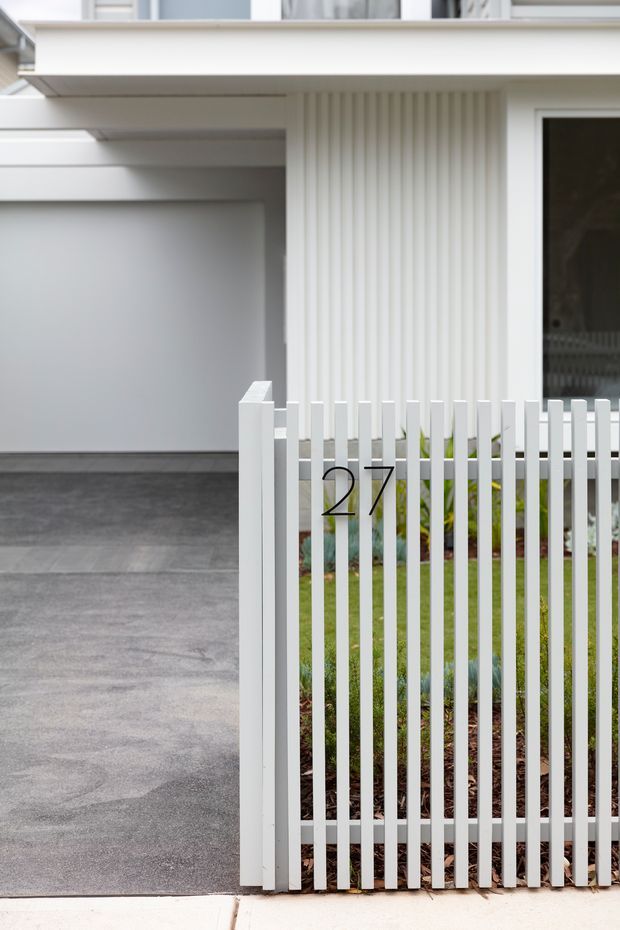Close-up exterior view of a contemporary white residential frontage, featuring a slim vertical picket fence and gate with the number 27, a small landscaped front garden, grey concrete driveway, covered entry or garage opening, and vertically ribbed white facade cladding.