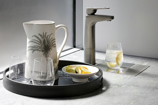 Close-up of a contemporary bathroom vanity area featuring a chrome single-lever tap, integrated linear drain, and grey stone or concrete-look countertop. A black tray with a decorative jug, glasses, and citrus sits in the foreground.