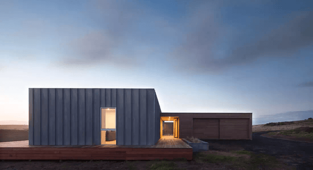 A minimalist, single-storey modern building set in a stark open landscape at dusk. The composition features a dark vertically ribbed metal-clad main volume with a narrow illuminated entry, connected to a lower dark garage volume. A broad timber deck runs along the front, with warm interior lighting contrasting against the cool blue-grey sky and dark terrain.