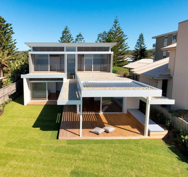 A bright exterior view of a contemporary multi-level home with extensive glazing, white rendered or painted structural elements, corrugated roof forms, and a generous timber deck beneath a covered outdoor area. The house sits within a landscaped suburban garden with mature conifer trees and neighbouring residences visible around it.
