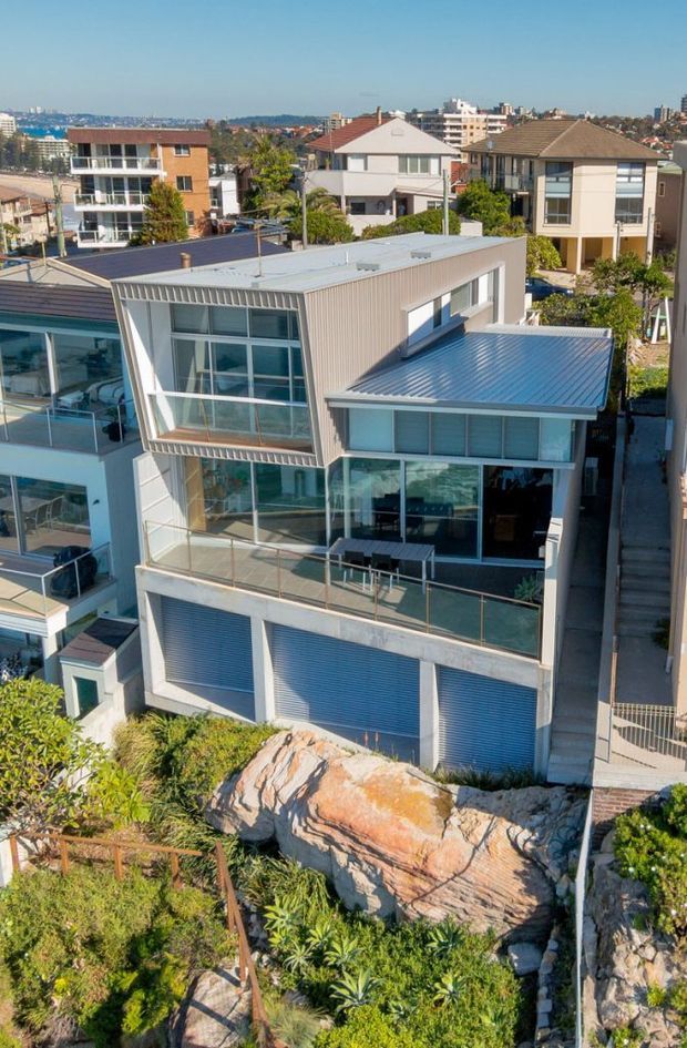 Elevated exterior view of a contemporary multi-level residential house on a sloping rocky site. The home features extensive floor-to-ceiling glazing, projecting balconies with glass balustrades, pale metal cladding, and corrugated metal roofs, with multiple garage doors at the lower level. Established neighbouring homes and an urban coastal-looking setting are visible in the background.