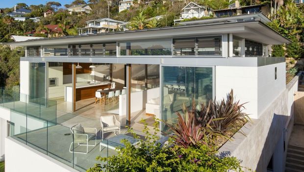 Contemporary multi-level home on a steep landscaped site, photographed from outside. The house features a low-pitched roof with broad eaves, expansive floor-to-ceiling glazing, white rendered walls, dark window frames, and a glass-balustraded terrace. An open-plan kitchen and dining area is visible through the glazing, along with an outdoor seating terrace and stairs descending along the side of the property.