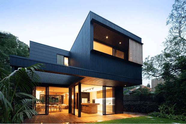 Exterior view of a contemporary two-storey residence with a dramatic steeply pitched roof form, dark vertical cladding, broad glazing, and warm illuminated interiors. The house opens onto a timber deck and landscaped garden, with a projecting upper-level volume and covered outdoor entertaining area.