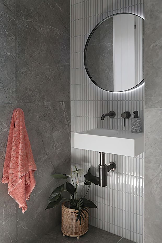 A compact contemporary bathroom with a wall-mounted white basin, black tapware and plumbing, a large round backlit mirror, grey stone-look wall tiles, and a vertical white fluted tile feature wall. A coral towel and potted plant add colour and texture.