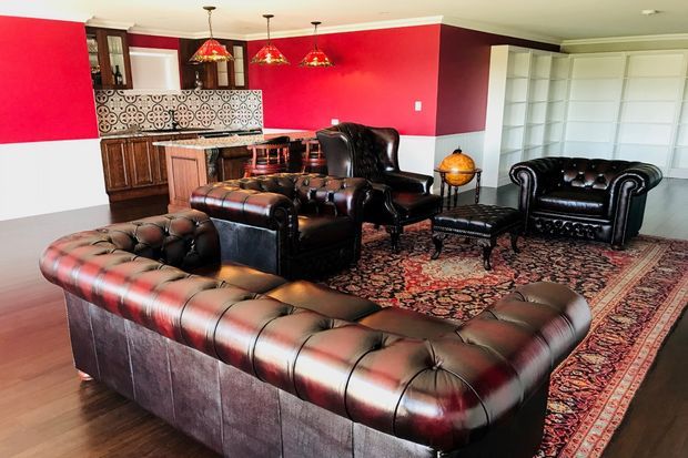 A richly styled living room with tufted dark leather Chesterfield seating arranged around a patterned Persian-style rug. The room features bold red and white walls, dark timber cabinetry, a decorative patterned backsplash, pendant lights, a globe bar cart, and built-in white storage.