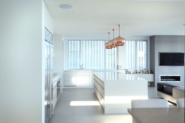 Bright, open-plan contemporary kitchen and living area with glossy white cabinetry, a large white island, pale tiled flooring, floor-to-ceiling vertical blinds, and copper pendant lights. A minimalist living zone with wall-mounted television and linear fireplace is visible beyond the kitchen.