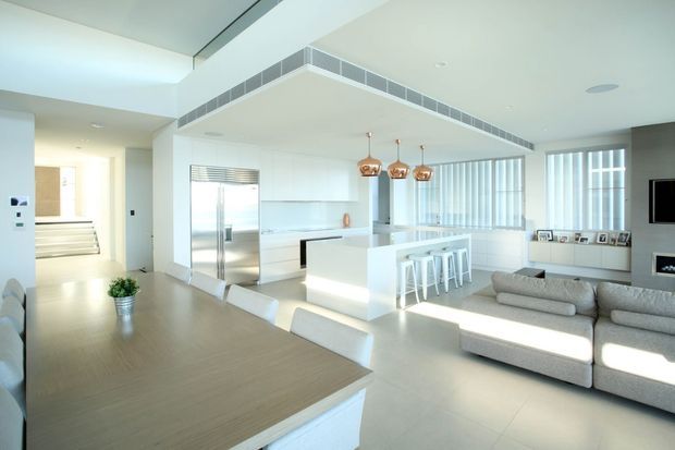 Bright, open-plan contemporary interior combining a white kitchen, dining area, and living room. The kitchen features a large island with white cabinetry and bar seating, stainless-steel appliances, and a tiled rangehood surround. A timber dining table occupies the foreground, while a large pale sectional sofa faces a wall-mounted television and fireplace. Floor-to-ceiling windows with vertical blinds provide abundant natural light, and copper pendant lights add a warm accent.