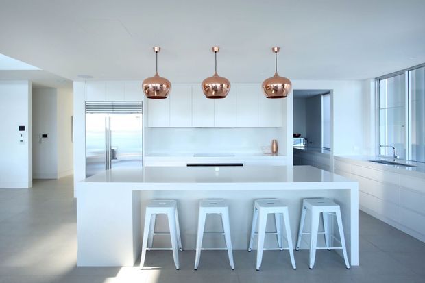Bright, spacious minimalist kitchen with a large white island, four white stools, integrated white cabinetry, stainless-steel refrigerator, and expansive glazing along the right side. Three copper pendant lights provide warm accents above the island, while pale flooring and surfaces create a clean, contemporary interior.