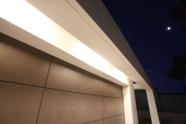 Night-time close-up of a contemporary building facade with large warm-toned rectangular wall panels, crisp white horizontal soffit and trim, and a bright linear recessed light. A white structural frame surrounds dark glazing at the right, with a deep blue night sky and moon beyond.