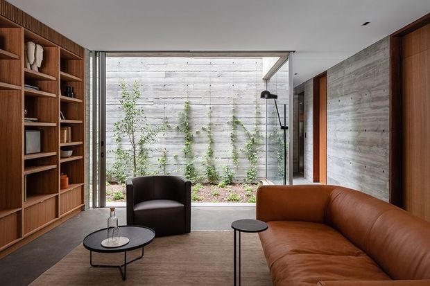 A refined contemporary living room with a large glazed opening to a planted courtyard. The room features extensive dark timber built-in shelving, a grey concrete or stone-textured feature wall, polished concrete flooring, a brown leather sectional sofa, a black lounge chair, and minimalist black side tables and floor lamp. The space has a calm, warm, indoor-outdoor character with strong horizontal lines and abundant natural light.