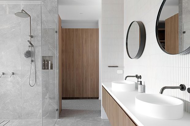 Contemporary bathroom with a glass-enclosed walk-in shower, pale marble-look wall and floor tiles, twin white vessel basins, a timber-veneer floating vanity, brushed dark metal tapware, and round black-framed mirrors. A timber-clad doorway leads toward an adjacent private area.