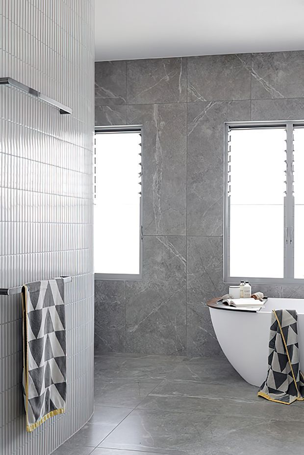 A bright contemporary bathroom featuring a freestanding white bathtub, large-format grey marble-look wall and floor tiles, and a curved wall clad in narrow white vertical tiles. Two windows provide abundant natural light, while monochrome geometric towels add a small decorative accent.