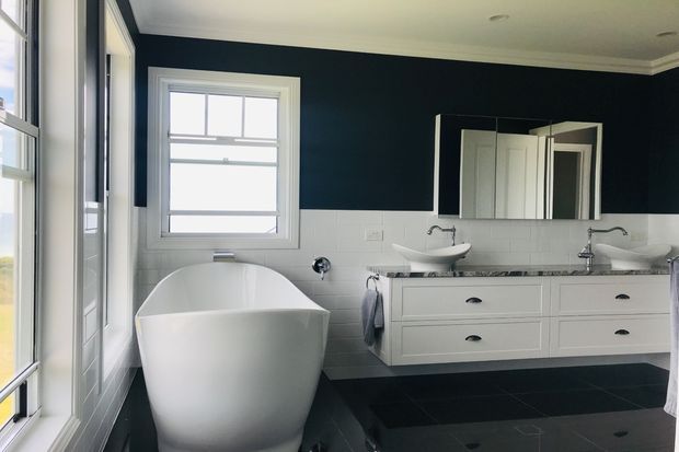 A bright, refined bathroom featuring a freestanding white bathtub beside large windows, a long double-sink vanity with white cabinetry, wall-mounted mirrors, chrome fixtures, white tiled lower walls, dark navy upper walls, and dark floor tiles.