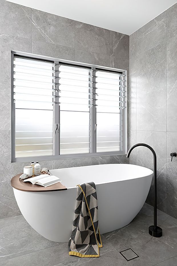 A bright contemporary bathroom featuring a freestanding white oval bathtub, grey marble-look wall and floor tiles, a black floor-mounted bath filler, and a large aluminium-framed louvred window. A curved timber bath tray with books and toiletries, plus a patterned towel, add warmth and texture.