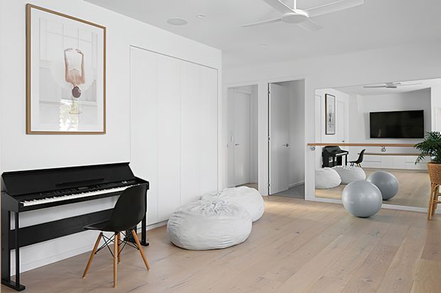 Bright, minimalist open-plan interior with pale timber flooring, white walls and built-in cabinetry. A black upright piano and framed artwork sit along the left wall, with white beanbags in the central area. Through the wide opening and mirror, a living area with a television, low white joinery, desk, chair and exercise balls is visible. A ceiling fan and indoor plant add functional and natural accents.