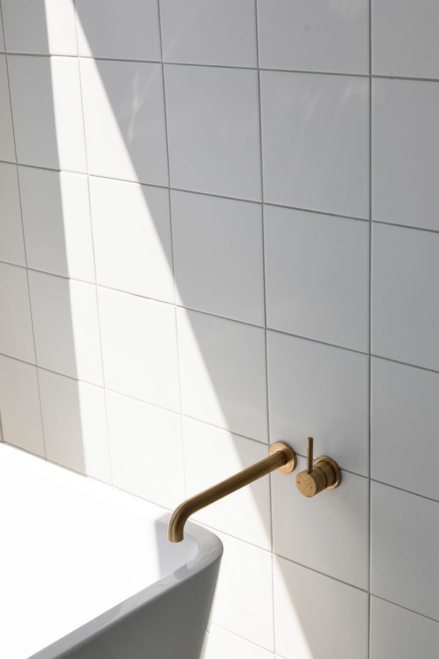 Close-up of a minimalist bathroom with a white freestanding bathtub partially visible against a wall of large white square tiles. A wall-mounted brushed brass tub filler and matching control are featured, with strong sunlight casting geometric shadows across the tiled surface.