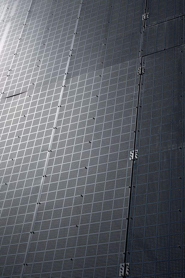 Close-up view of a tall, dark grey perforated metal building facade composed of repeating square-grid panels. Vertical panel joints and exposed fastening brackets are visible, with directional lighting creating a subtle gradient across the surface. The image focuses on the exterior cladding and its industrial geometric texture.