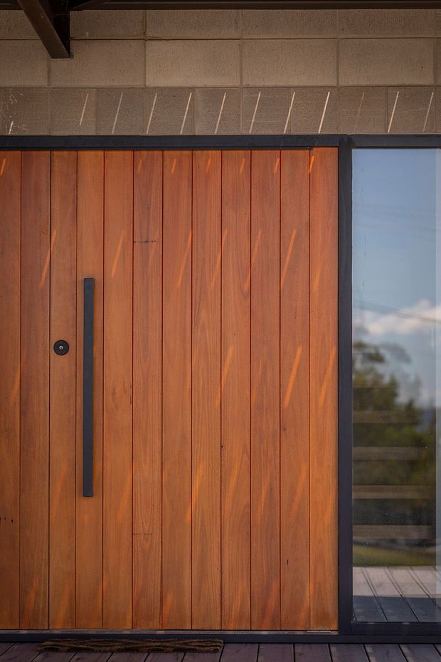 Close-up exterior view of a contemporary entry door clad in vertically arranged warm timber boards, with a long matte-black pull handle and matching dark frame. A narrow glazed sidelight reflects the surrounding landscape, while the upper facade features textured grey masonry or concrete blockwork. Dark paving and a doormat sit at the threshold.