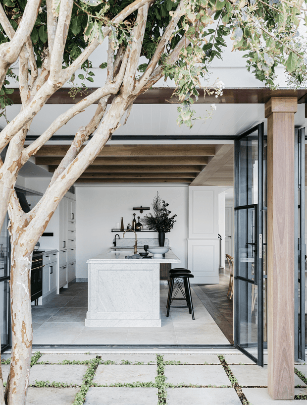 A bright, refined kitchen viewed from an outdoor paved courtyard through fully open black-framed glazed doors. The space features a large white marble island with a brass tap and black stools, white shaker-style cabinetry, dark tiled flooring, timber ceiling beams, and a white paneled door. A mature tree frames the foreground and a timber-clad structural post defines the opening.