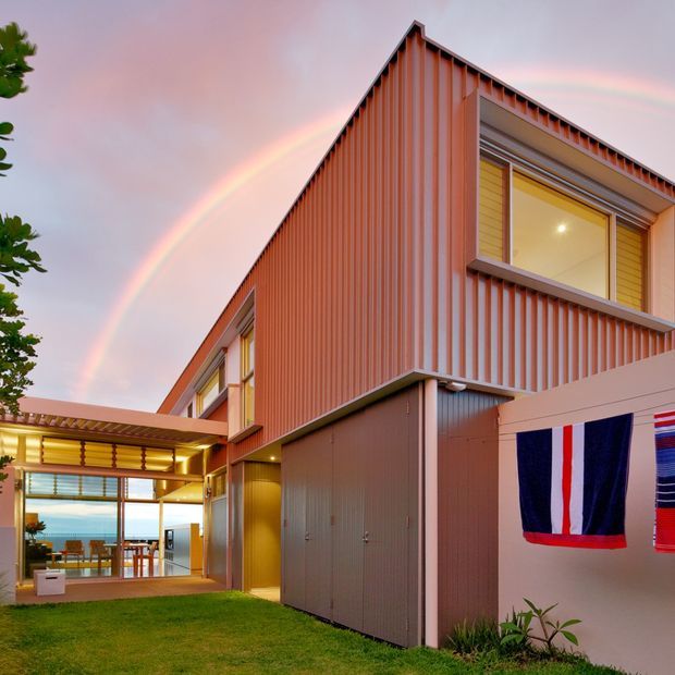 Exterior view of a contemporary multi-level home with a sharply pitched gable-like roof form and vertically ribbed warm terracotta metal cladding. Large windows and an illuminated glazed living or dining area open onto a small lawn courtyard, with coastal water visible in the distance. A rainbow and hanging textile appear in the scene.