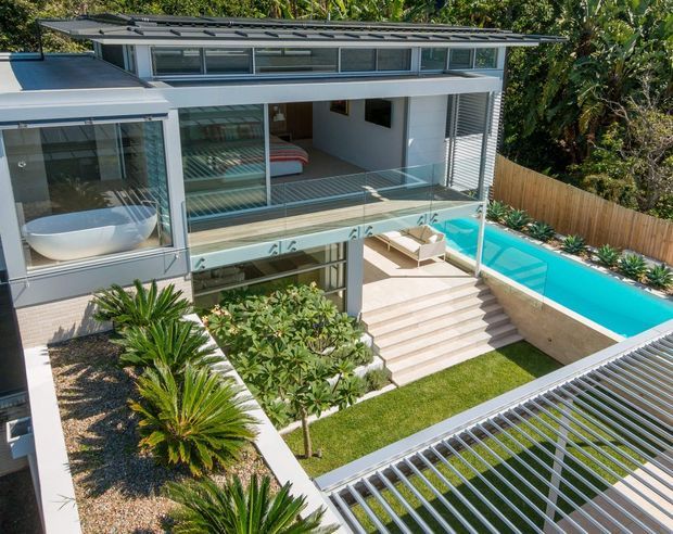 Aerial view of a contemporary multi-level residence with extensive glazing, slim metal roof forms, a covered outdoor terrace, and a bright turquoise swimming pool. The house is arranged around landscaped garden areas with palms, gravel beds, lawn, and timber screening. A large slatted structure occupies the foreground, likely a carport or covered outdoor platform.