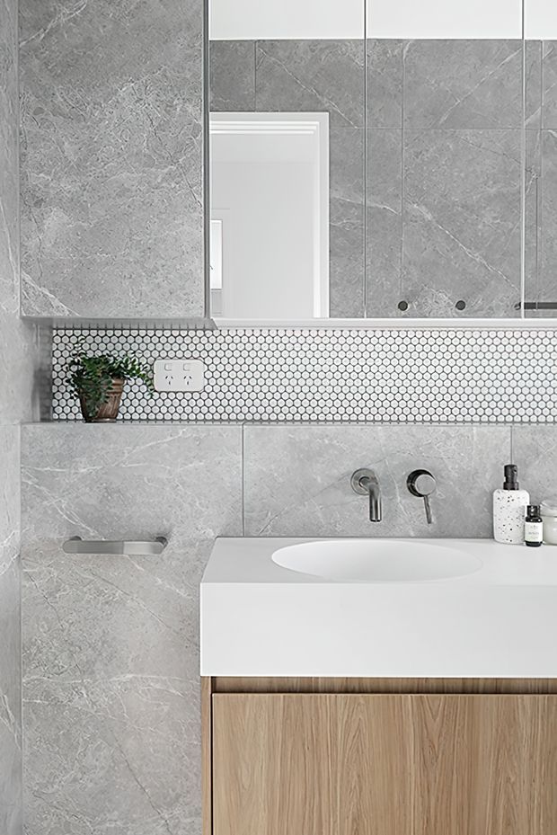 Contemporary bathroom vanity area featuring large grey marble-look wall tiles, a white integrated basin and benchtop, timber-look vanity cabinetry, and a narrow white-and-grey mosaic tile backsplash beneath a mirrored cabinet. Brushed metal wall-mounted tapware and minimal accessories complete the clean, refined scheme.